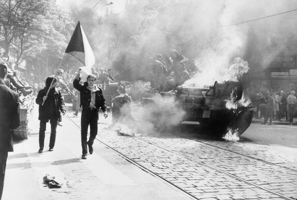 21. srpen 1968 v Československu – obyvatelé se bouří proti okupaci, v pozadí hoří sovětský tank.